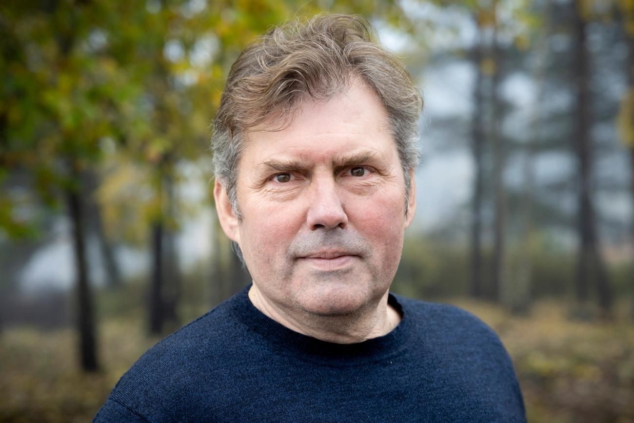 Man in a black sweater standing outdoors with a blurred tree background.