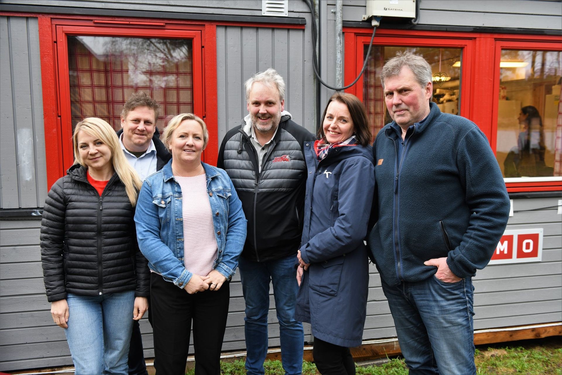 Group of people standing outside in front of a grey building with red window frames.