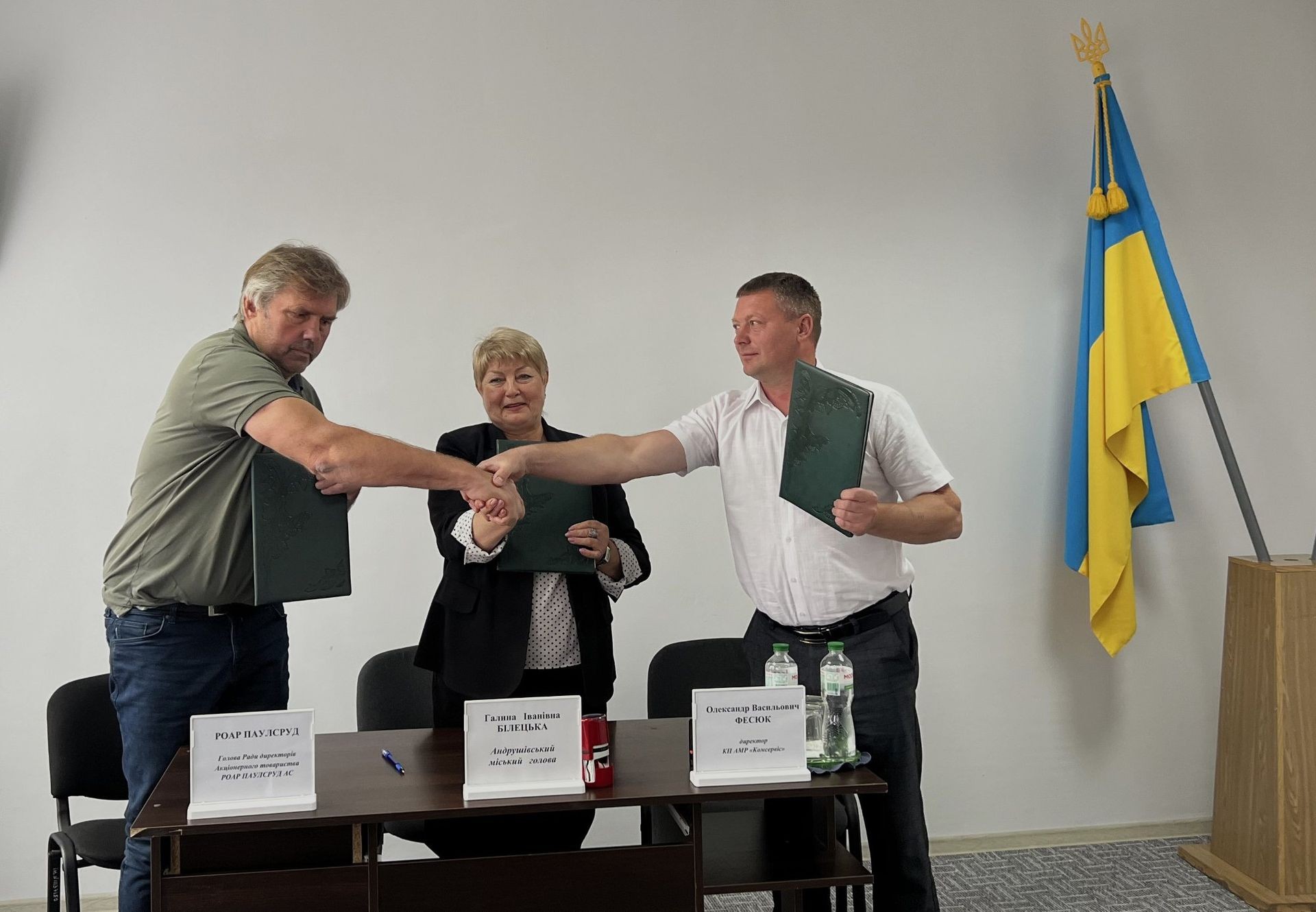 Three people shaking hands at a table with folders, next to a Ukrainian flag.